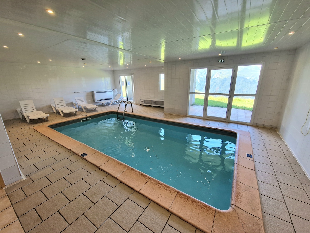 An indoor swimming pool is featured, surrounded by tiled flooring and illuminated by natural light from large windows. Lounge chairs are neatly arranged along one side, while access to a scenic outdoor area is available through glass doors.