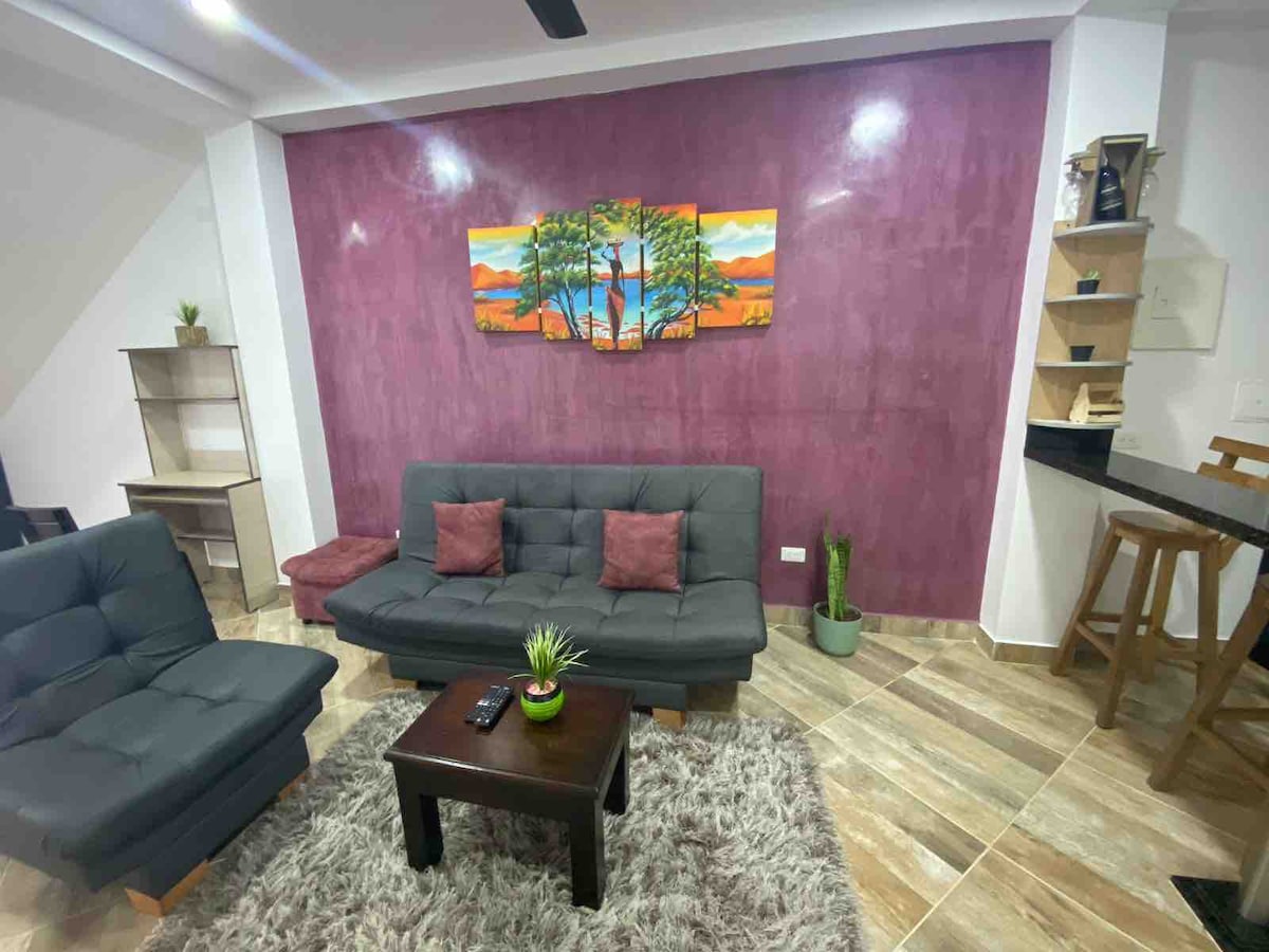A modern living area features a dark purple accent wall adorned with an artistic landscape painting. Comfortable seating comprises a dark sofa and armchairs against a textured rug. A small table and potted plant add touches of comfort, while a minimal bar area is visible in the background.