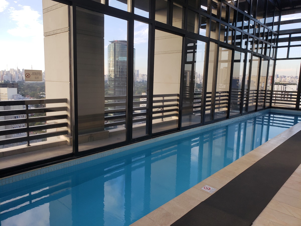 An indoor pool, framed by large glass windows, offers a clear view of the skyline. The pool features calm blue water and is bordered by a tiled deck. Natural light fills the space, enhancing the serene atmosphere.