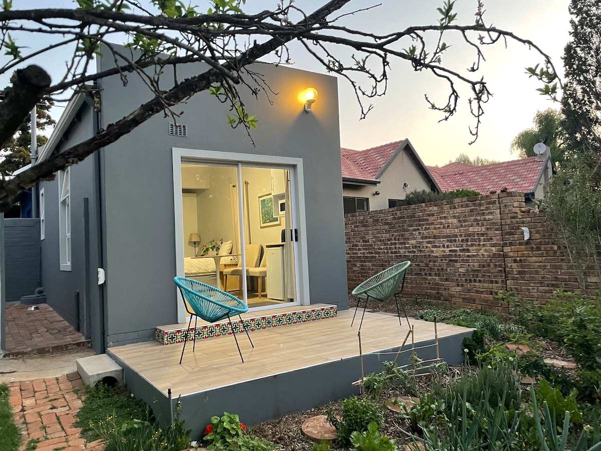The exterior of a garden cottage is depicted, featuring a wooden deck with two stylish chairs. A welcoming entrance opens into the interior, which is partially visible. Lush greenery and garden elements surround the cottage, creating a serene outdoor setting.