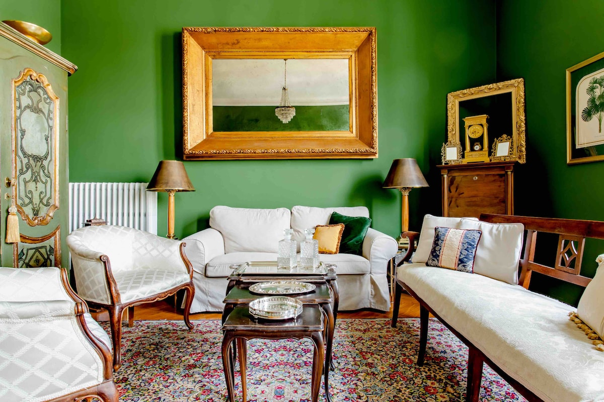 A living area features a deep green wall, framed by classic furniture pieces. A large mirror hangs above a white sofa, complemented by two elegant armchairs. A low wooden table with decorative items sits at the center, while traditional artwork and a chandelier add a touch of sophistication.