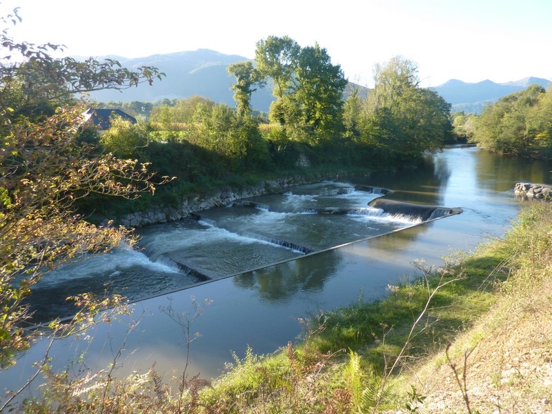 A serene river flows gently, with a series of small cascading waterfalls visible. Lush greenery lines both banks, and distant mountains can be seen under a clear sky. The tranquil scene reflects a natural landscape, inviting exploration and relaxation.