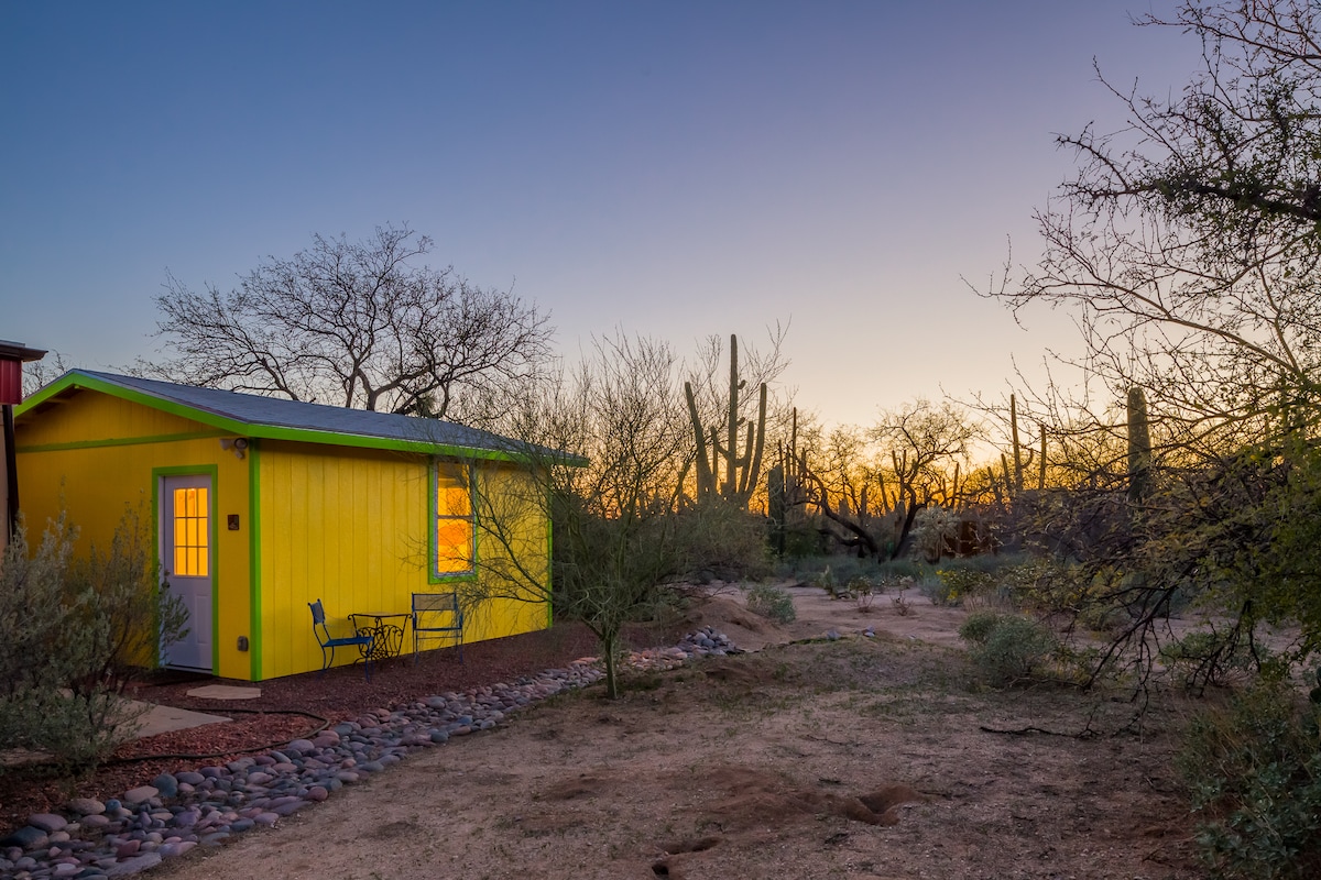 A vibrant yellow casita stands amid desert vegetation at dusk, with a soft glow emanating from the windows. Silhouettes of tall cacti are visible in the background against a gradient sky transitioning from orange to deep blue.