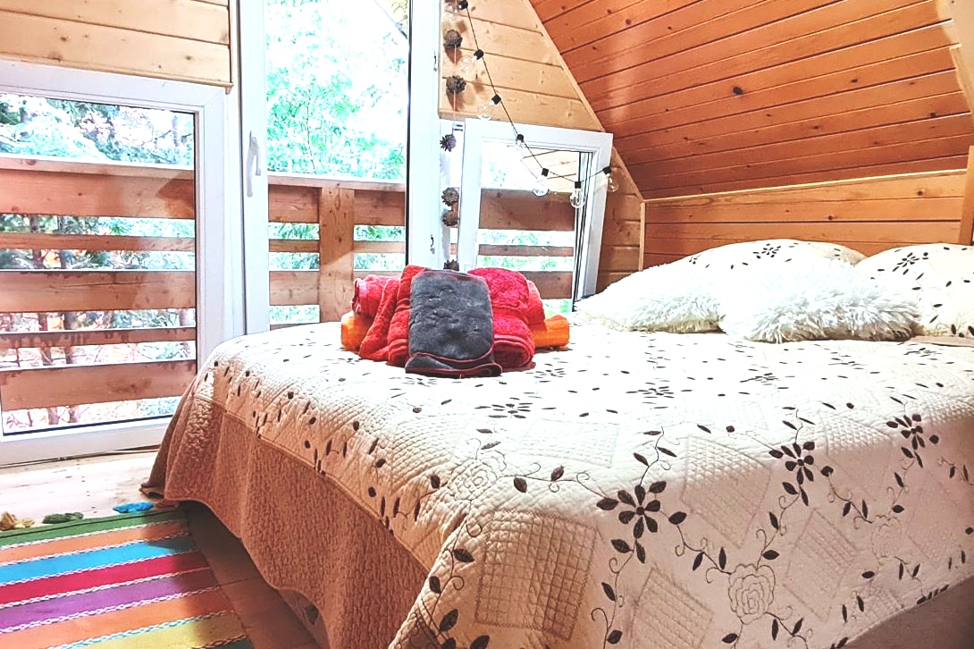 A spacious sleeping area is highlighted by a quilted bedspread featuring floral patterns. Two fluffy pillows and towels are arranged on the bed. Large windows offer views of the greenery outside, while a colorful woven rug adds warmth to the wooden floor.
