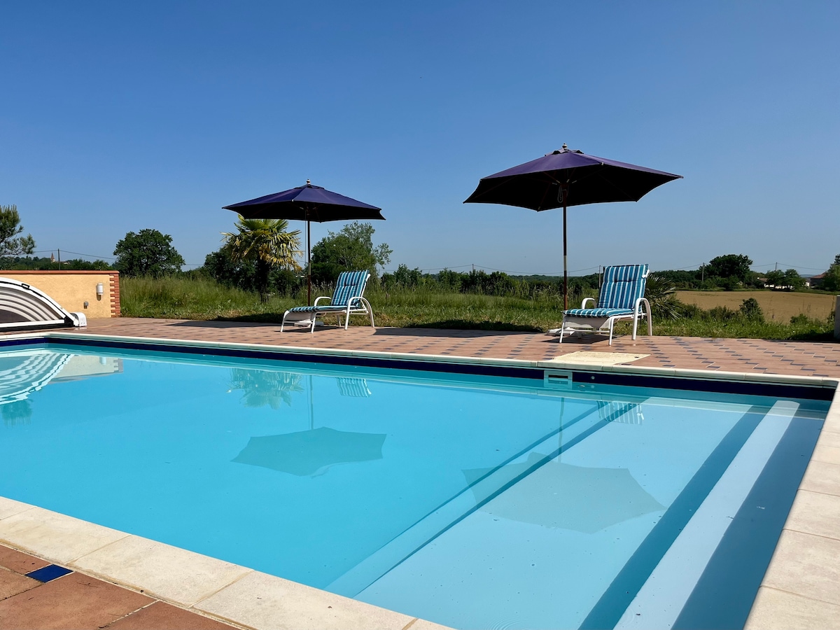 A clear blue swimming pool is surrounded by a sun patio, featuring two striped lounge chairs under expansive umbrellas. Lush greenery and open fields are visible in the background under a bright blue sky.