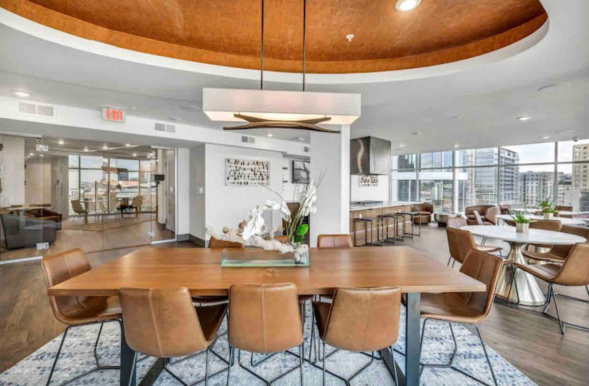 An expansive dining area features a large, wooden table surrounded by brown leather chairs. Natural light fills the space through large windows, highlighting a modern chandelier overhead. A patterned rug adds texture to the floor, complementing the open layout that includes a glimpse of a sleek kitchen.