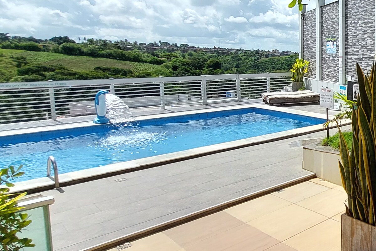 An outdoor swimming pool is set against a backdrop of green hills, with sunlight reflecting off the water's surface. A poolside lounge area with a sunbed is visible nearby. Lush plants are placed around the pool area, enhancing the serene environment.
