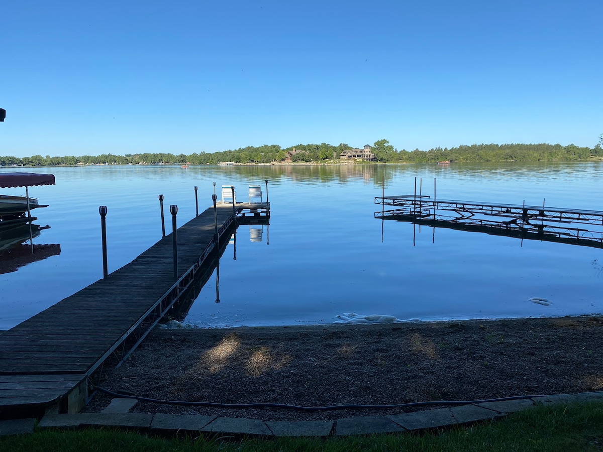 A serene view of Pickerel Lake features calm waters reflecting the clear blue sky. A wooden dock extends into the lake, accompanied by two boat slips. The gentle shoreline is lined with grass, creating a peaceful atmosphere for outdoor activities.