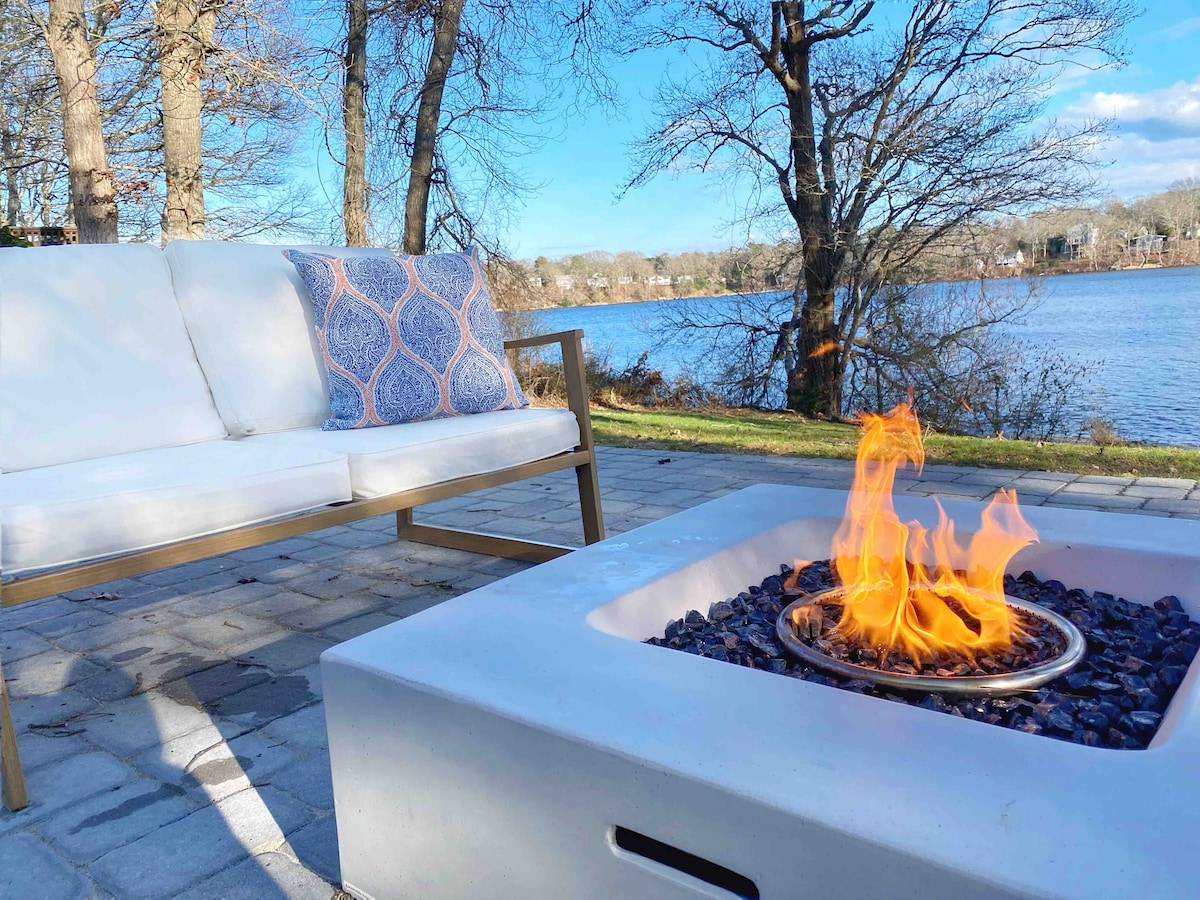 A modern outdoor seating area is displayed near Jenkins Pond, featuring a white sofa with patterned cushions. A fire pit with a flame burns in the foreground, surrounded by decorative stones. Leafless trees frame the tranquil water view in the background.