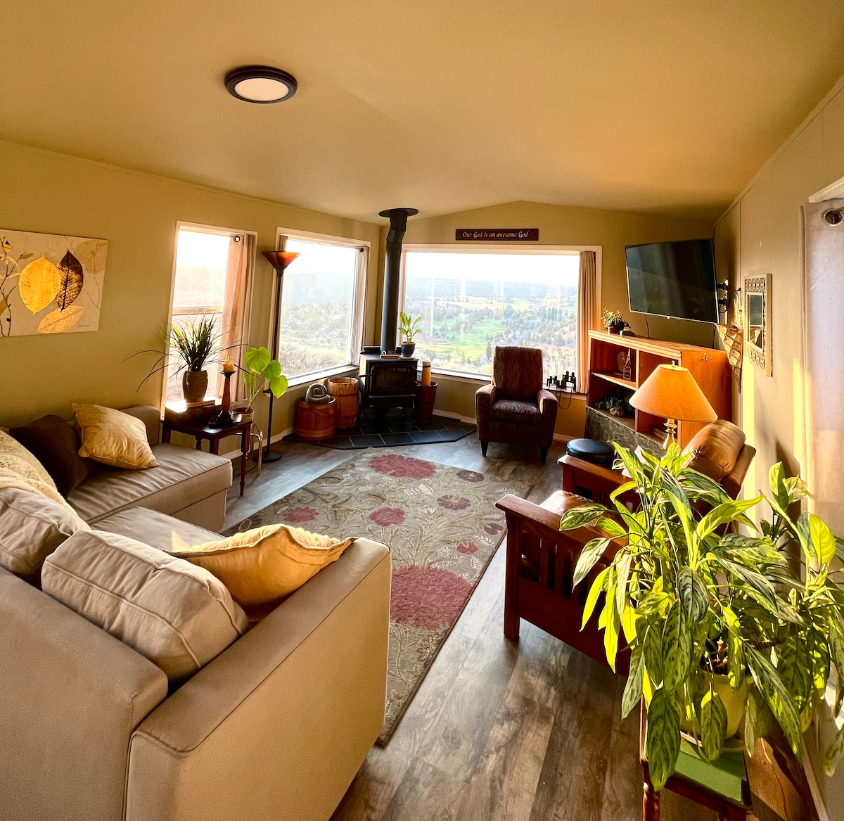 A bright living area features large windows offering expansive views of the valley. Comfortable seating includes a spacious couch, an armchair, and a wooden coffee table. A wood stove adds warmth, while plants and artwork provide natural decor elements. A TV is mounted on the wall.