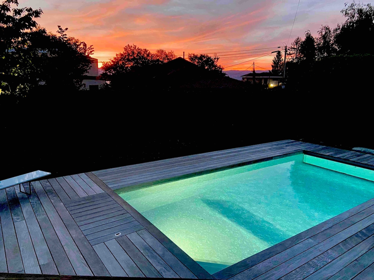 A warm evening scene features a heated pool set against a colorful sunset. The pool is surrounded by wooden decking, with reflections of the vibrant sky visible on the water's surface. Surrounding trees and buildings outline the tranquil environment.