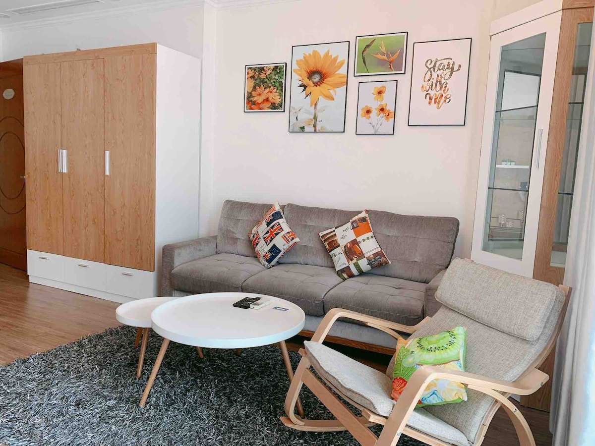 A cozy living room features a gray sofa adorned with colorful cushions, positioned beside a light wooden chair. A round white coffee table sits on a textured rug, while framed artwork of flowers and a motivational quote decorates the wall above. Storage cabinets are visible along one side.