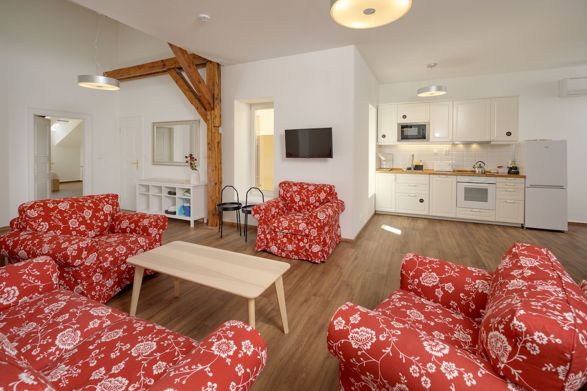 A welcoming living area is furnished with two red floral-patterned sofas and a light wooden coffee table. A flat-screen television is mounted on the wall, while a stylish kitchen with white cabinetry and modern appliances is visible in the background.
