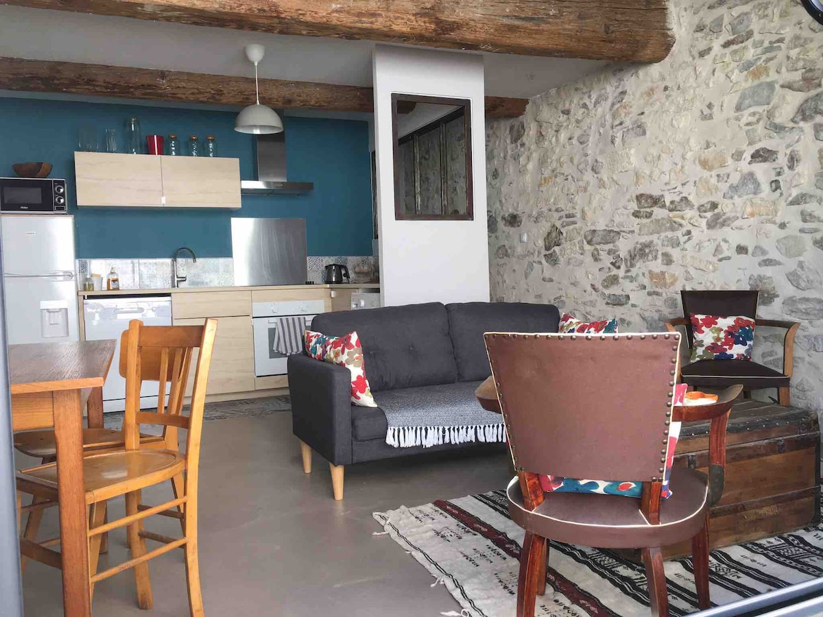 An open living space is present, featuring a modern kitchen with light cabinetry and a dining table surrounded by wooden chairs. A comfortable sofa is centered with decorative cushions, and a cozy area rug enhances the floor. Exposed stone walls add texture to the room.