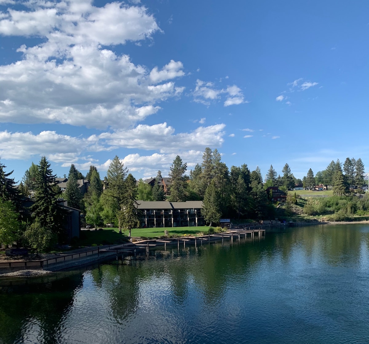 The image captures a serene view of the water, reflecting the blue sky and scattered clouds. Lush green trees frame the shoreline, while an array of buildings stands neatly along the waterfront. The vibrant landscape showcases the peaceful atmosphere of the surroundings.