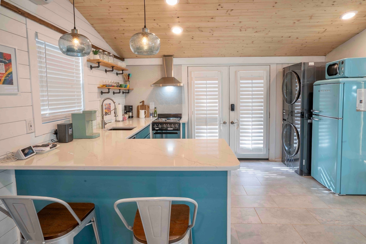 A bright kitchen area features modern appliances, including a blue refrigerator and stainless steel stove. A quartz countertop extends into an inviting bar with two metal stools. Double French doors lead to the outdoor space, while shelves display decorative items and kitchen essentials.