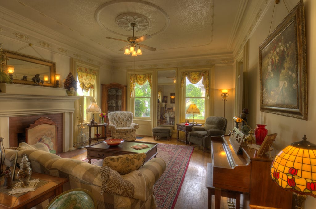 The living area features a mix of vintage furniture, including a checkered sofa and an upright piano. Warm light illuminates the space through large windows framed by floral curtains. Decorative items and a colorful rug add character to the room, which has intricate ceiling details.