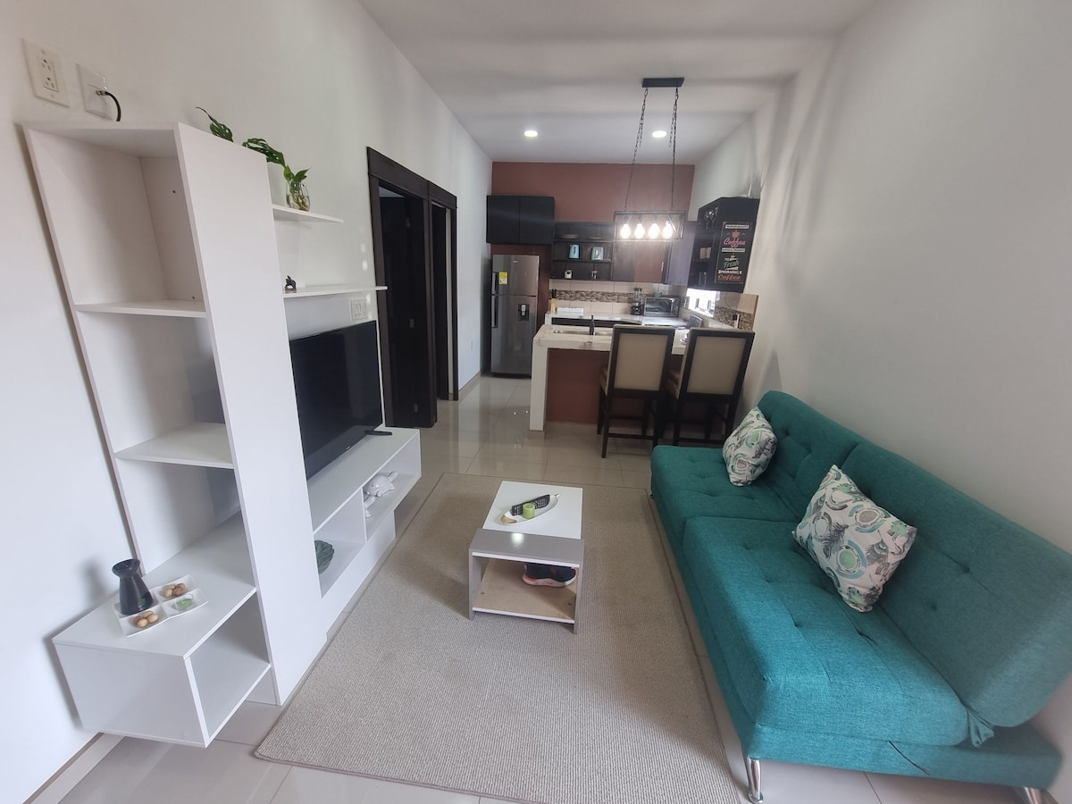 A living area is showcased, featuring a modern turquoise sofa and a small coffee table atop a neutral area rug. A wall-mounted television is positioned on the left, while the kitchen area is visible in the background, equipped with bar stools and appliances.