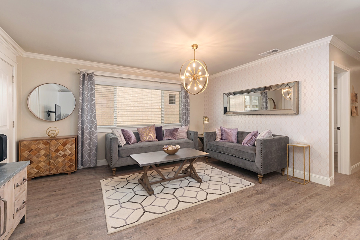 A stylish living room is showcased, featuring two gray sofas adorned with purple accent pillows. A wooden coffee table sits atop a geometric rug, while a round mirror and a large wall mirror add elegance. Natural light filters through the window, enhancing the inviting atmosphere.