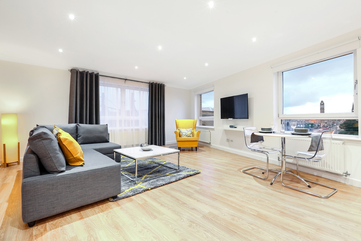 A modern living space features a gray sectional sofa and a bright yellow armchair. A flat-screen TV is mounted on the wall. Natural light floods the room through large windows, while a dining table with metal chairs is situated in one corner.