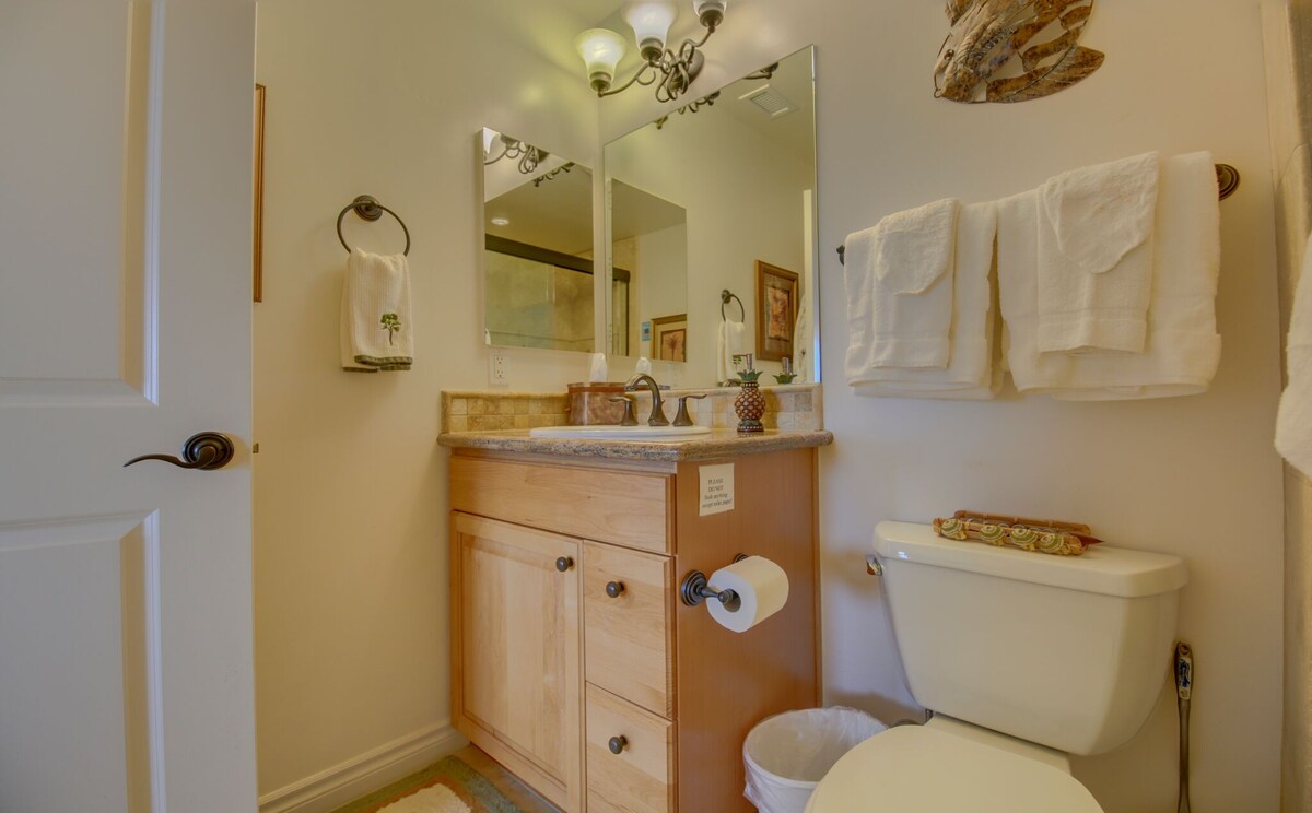 The bathroom features a wooden vanity with a granite countertop and dual mirrors above. Two towels are neatly hung on a rack, while additional towels are displayed nearby. A toilet and a wastebasket are present, contributing to a well-organized and functional space.