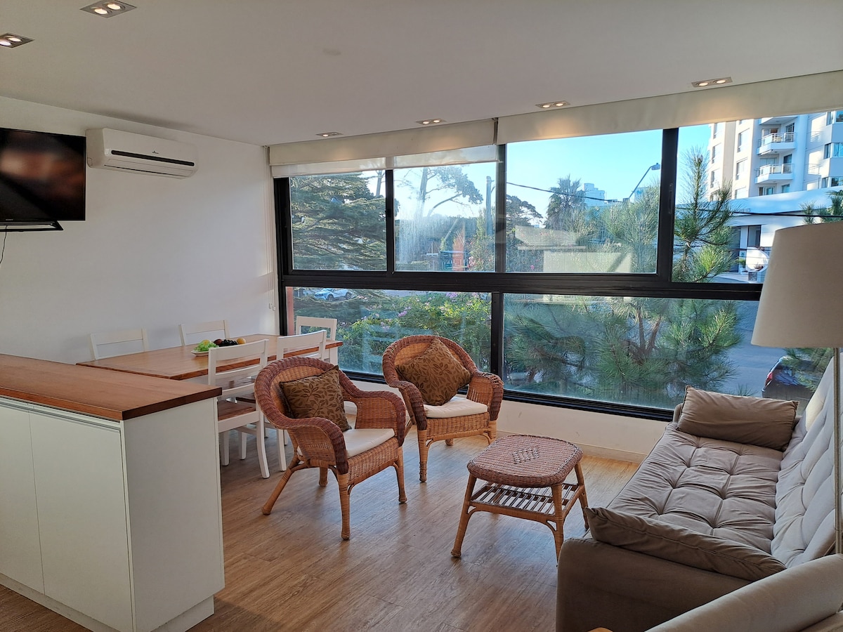 The living area features a comfortable sofa and two wicker chairs, arranged around a coffee table. Large windows provide natural light and offer views of the surrounding greenery. A dining table is positioned in the background, with an air conditioning unit mounted on the wall.