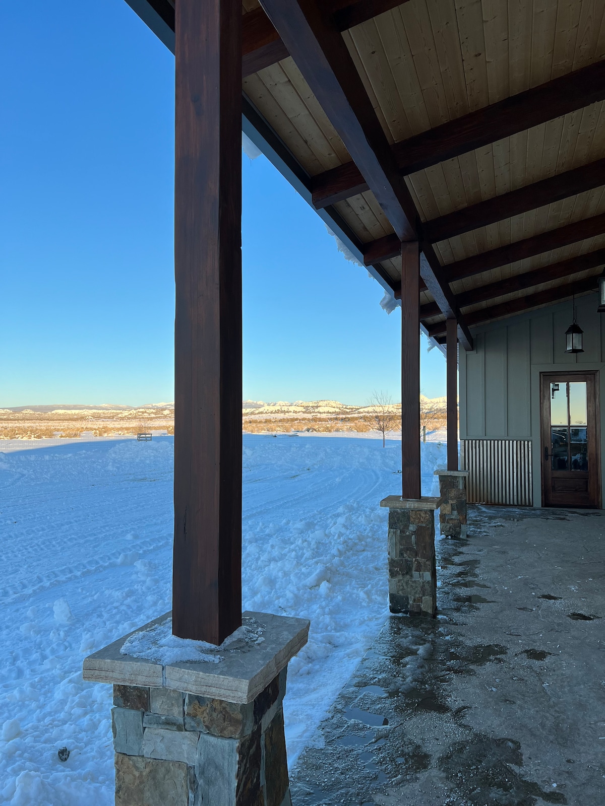 The exterior view features a covered porch supported by wooden beams, framed by stone pillars. A snowy landscape extends into the horizon, with distant mountains visible under a clear blue sky. The entrance to the interior space is seen to the right.