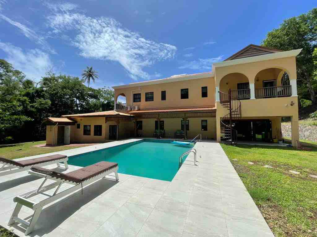 A spacious outdoor area is highlighted by a sparkling pool surrounded by lounge chairs. The two-story house features large arched windows, a shaded terrace, and a spiral staircase leading to the upper floor. Lush greenery frames the refreshing setting, with a clear blue sky above.