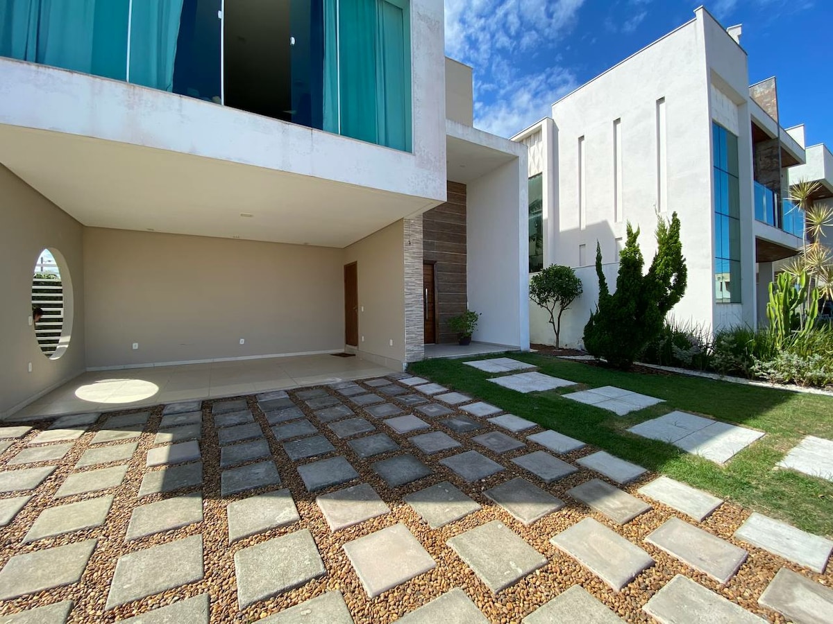 A spacious outdoor area is presented, featuring a patterned stone pathway leading toward a modern building. Lush greenery borders the space, complemented by a circular window that adds architectural interest. The bright blue sky enhances the inviting atmosphere of the property.