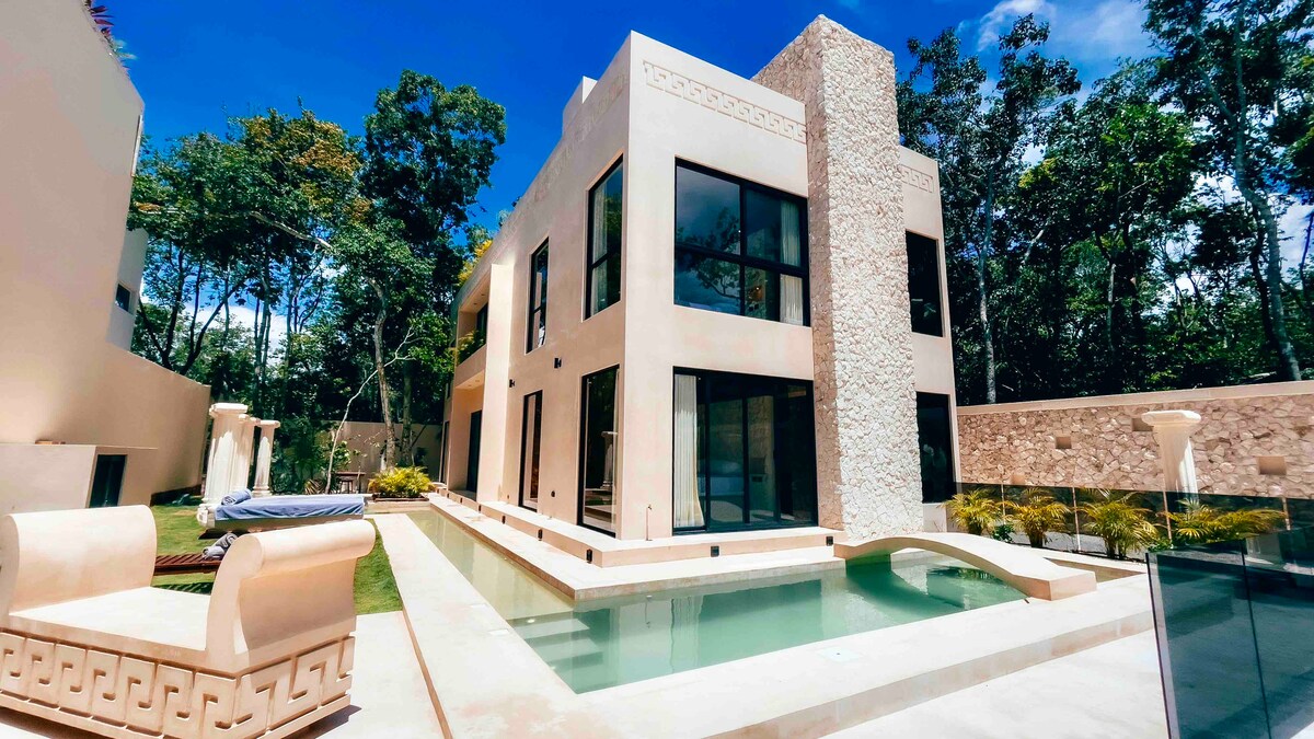 A modern villa is surrounded by lush greenery, featuring large glass windows that invite natural light. The exterior includes a smooth-stone facade and a serene swimming pool that reflects the vibrant sky. Comfortable outdoor seating is positioned near the pool area.