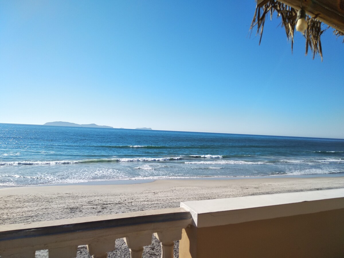 An expansive view of a tranquil beach is captured, showcasing gentle waves lapping at the shore. Clear blue skies and distant islands enhance the serene coastal landscape, framed by a balcony railing with rustic details.