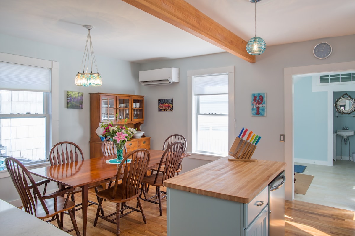 A well-lit dining area features a wooden table with six matching chairs, positioned next to a large window. A display cabinet holds decorative items. A light fixture with blue accents hangs above, while a wall-mounted air conditioner and clock add functionality to the bright space.