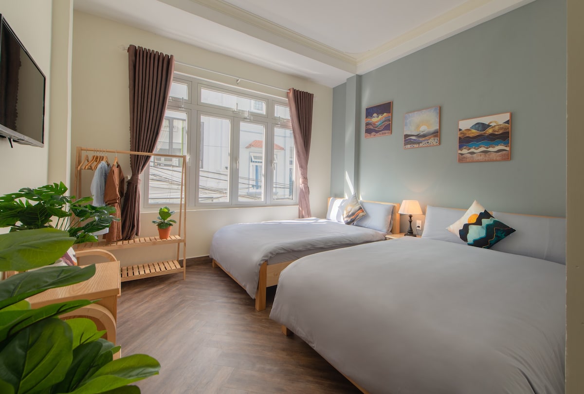 A bright sleeping area is presented, featuring two spacious beds with light grey linens. Natural light enters through large windows adorned with matching curtains. A wooden rack holds clothes, while a small plant adds a touch of greenery, enhancing the inviting environment.
