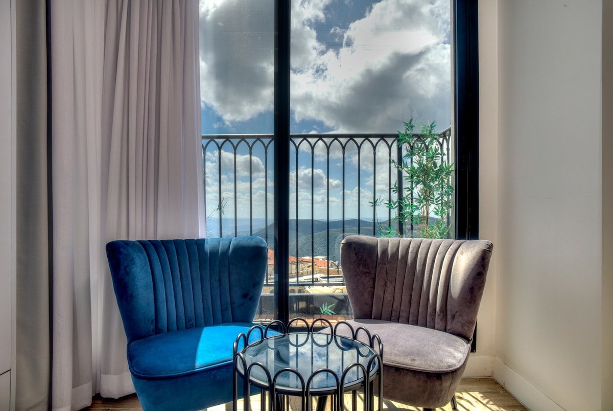 Two plush armchairs in complementary colors are positioned by large glass doors, offering a view of the outdoors. A small, round tabletop sits between the chairs, while soft curtains frame the scene, blending indoor comfort with natural light and the surrounding landscape.