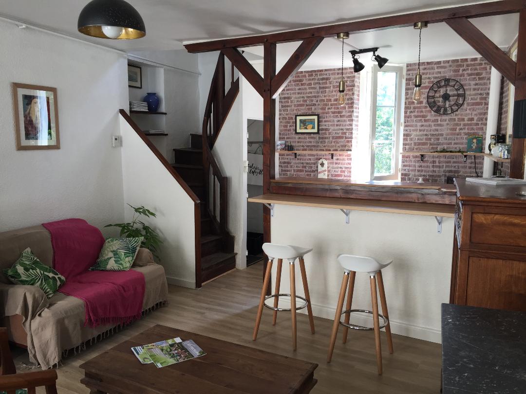 A bright living area features a cozy sofa draped with a pink throw, accompanied by a rustic wooden table. A bar with two white stools overlooks the kitchen area, which is highlighted by exposed brick walls and wooden beams. A staircase leads to an upper level.