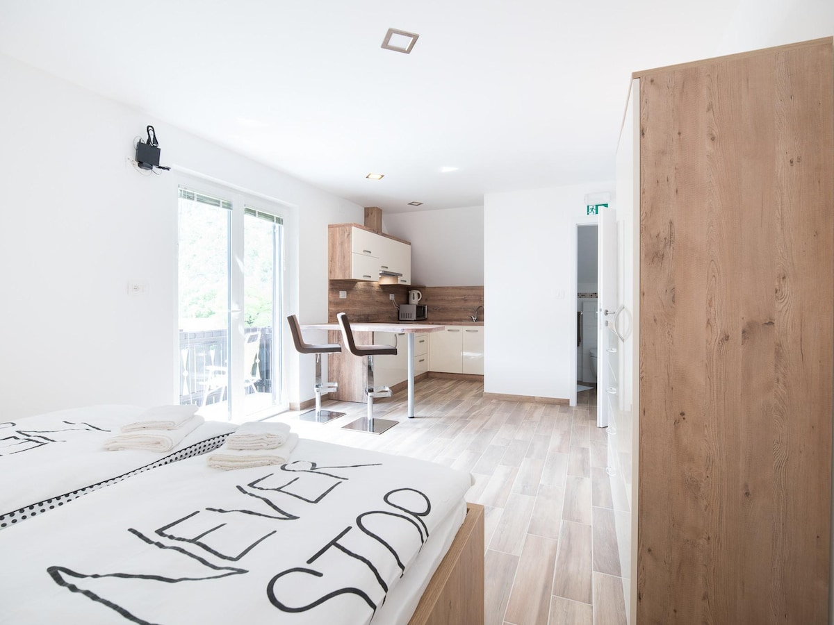 A modern apartment is displayed with light-colored wooden flooring and bright white walls. A spacious bed is positioned in the foreground, featuring a large decorative blanket. A kitchenette with contemporary appliances is visible in the background, alongside a dining area with two chairs and a small table.