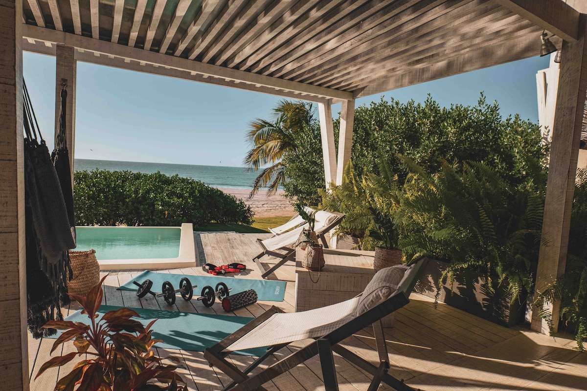 A serene patio area features comfortable lounge chairs and a refreshing pool, overlooking the beach. Lush greenery and decorative plants surround the space, with ocean views visible in the background. Fitness equipment rests nearby, enhancing the area for relaxation and activity.