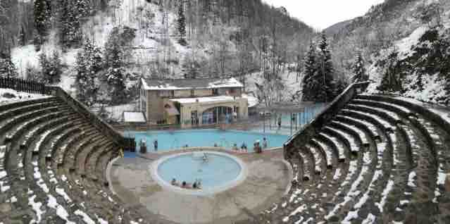 An outdoor pool area is set against a snowy mountainous backdrop, featuring tiered seating made of stone. The pool is visible with guests enjoying the water, while the surrounding landscape is blanketed in white snow. A building is located in the background.