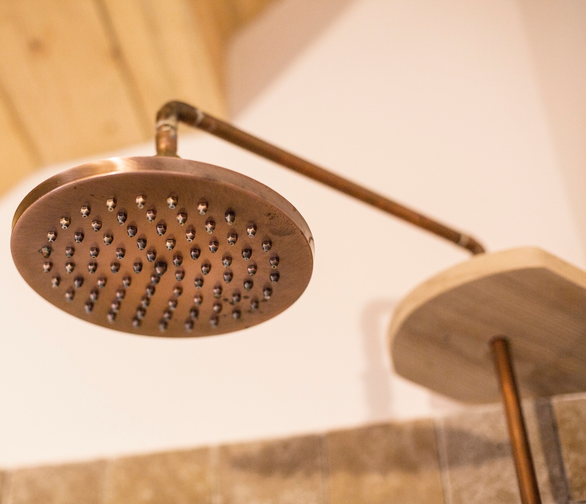 A copper showerhead is mounted on a sleek pipe, featuring numerous small nozzles arranged evenly across its surface. The warm metal finish complements the wooden elements in the background, creating a harmonious design in the bathroom space.
