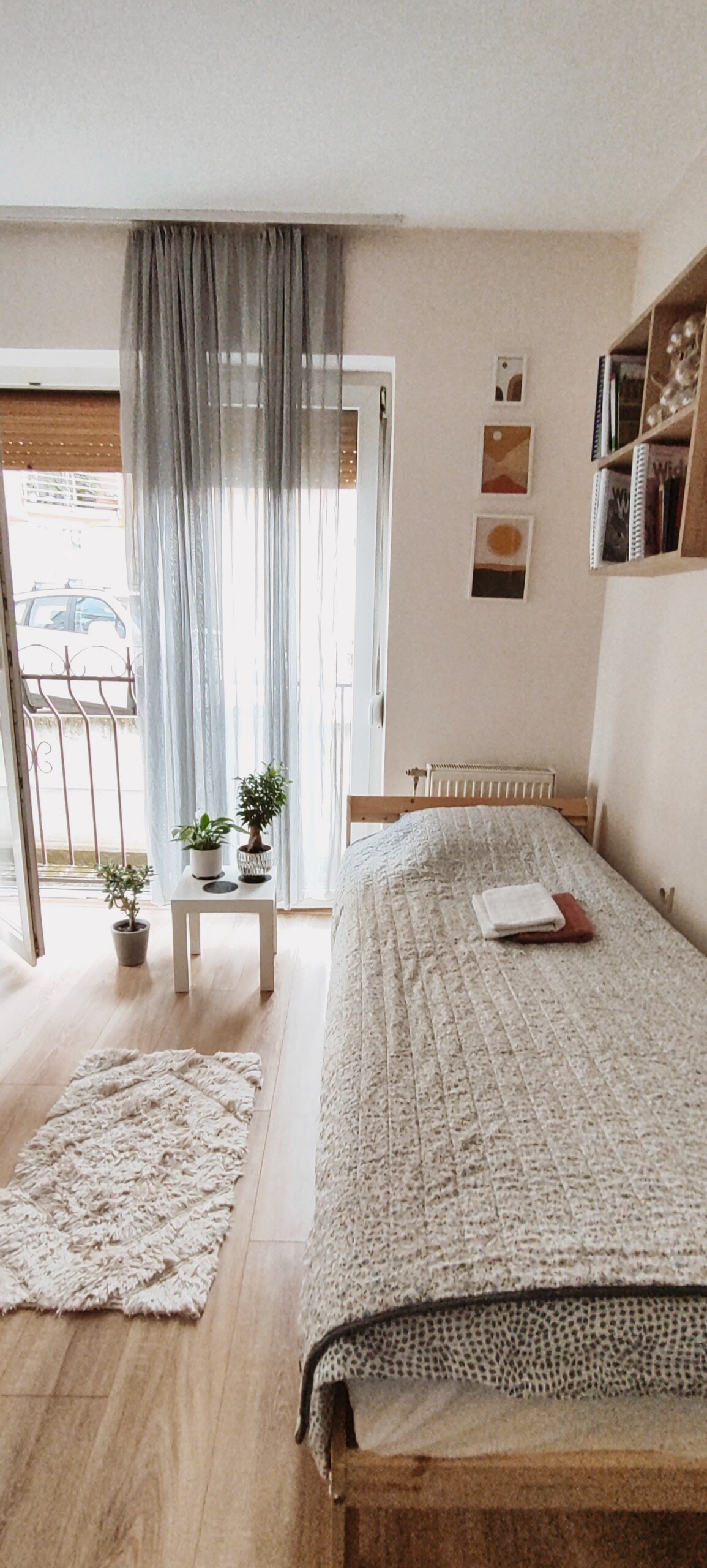 A cozy room is filled with natural light from a sliding glass door leading to a small balcony. A single bed is dressed in a soft blanket, with a book and a small plant on a side table. Decorative art pieces adorn the wall above a small bookshelf.