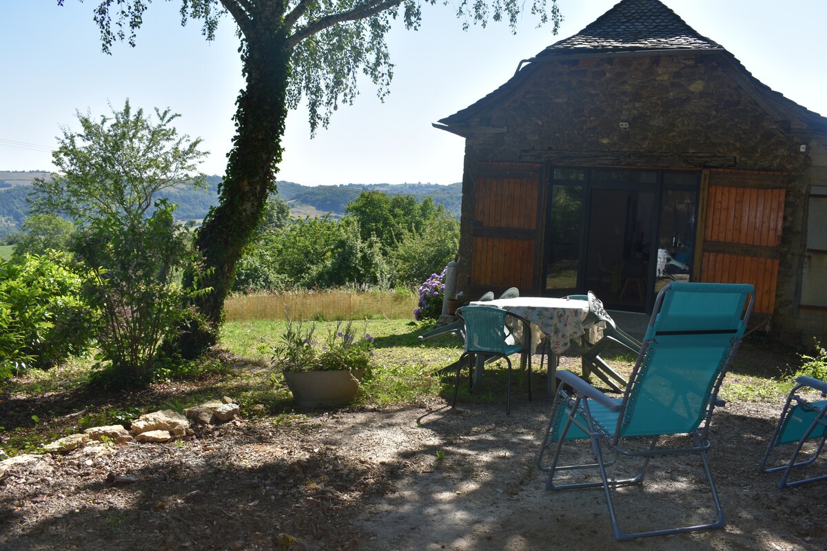 A charming exterior area features a small, round table with four chairs, situated under a tree. The backdrop displays a lush landscape and distant hills, enhancing the serene ambiance of the rustic stone building. A few potted plants add greenery to the scene.