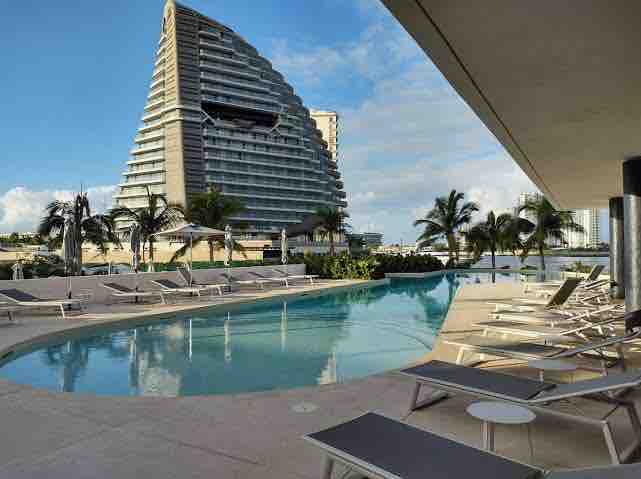 A tranquil pool area is bordered by lounge chairs, offering a serene atmosphere for relaxation. Lush greenery and palm trees are visible in the background, with a modern architectural structure rising in the distance against a clear blue sky.