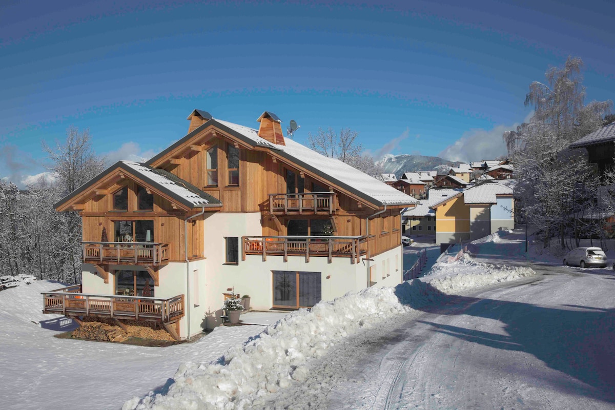 The exterior of a spacious chalet is shown, featuring wooden balconies and a sloped roof. A snowy landscape surrounds the building, with clear blue skies above. The pathway is partially cleared of snow, leading to the entrance, while nearby vehicles are parked along the road.