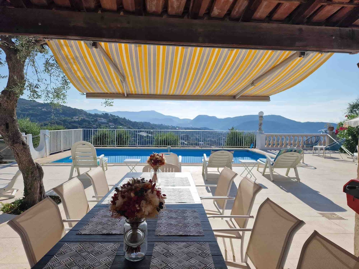 An outdoor dining area is presented with a large table set for meals, surrounded by several light-toned chairs. In the background, a spacious pool overlooks distant mountains, with an awning providing shade. The scene reflects a serene outdoor living space.