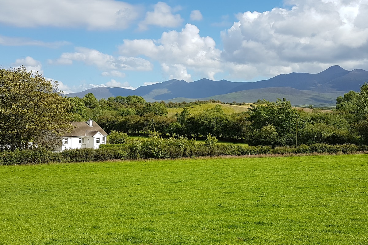 A charming white cottage is set against a backdrop of rolling green fields and distant mountains. Lush greenery surrounds the property, and a clear blue sky with fluffy clouds adds to the serene landscape. The peaceful countryside setting invites relaxation and exploration.