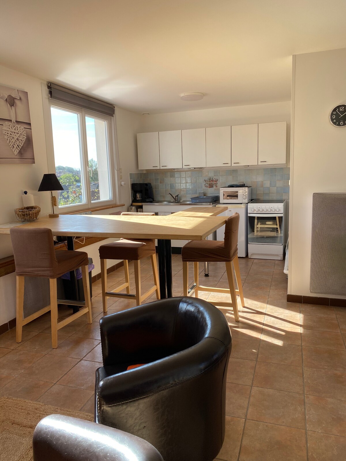 A bright kitchen area features light cabinetry and a spacious countertop with seating for four. Natural light fills the space through a window, illuminating the tiled floor and various appliances, including a coffee maker and oven, positioned along the back wall.