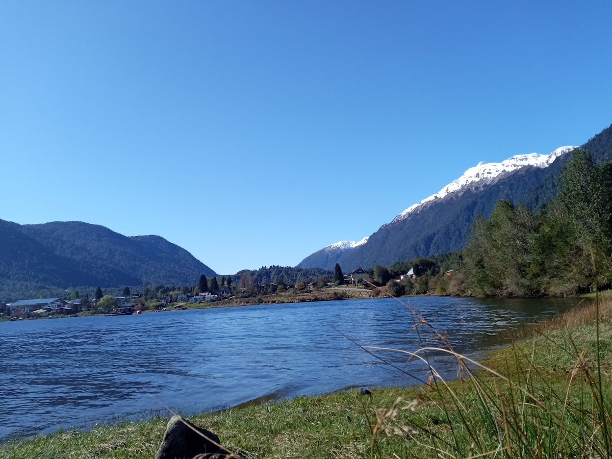 A serene view of a lake bordered by rolling hills and mountains. The landscape features a clear blue sky, snow-capped peaks in the distance, and lush greenery along the water's edge. Gentle ripples are visible on the lake's surface, enhancing the tranquil scene.