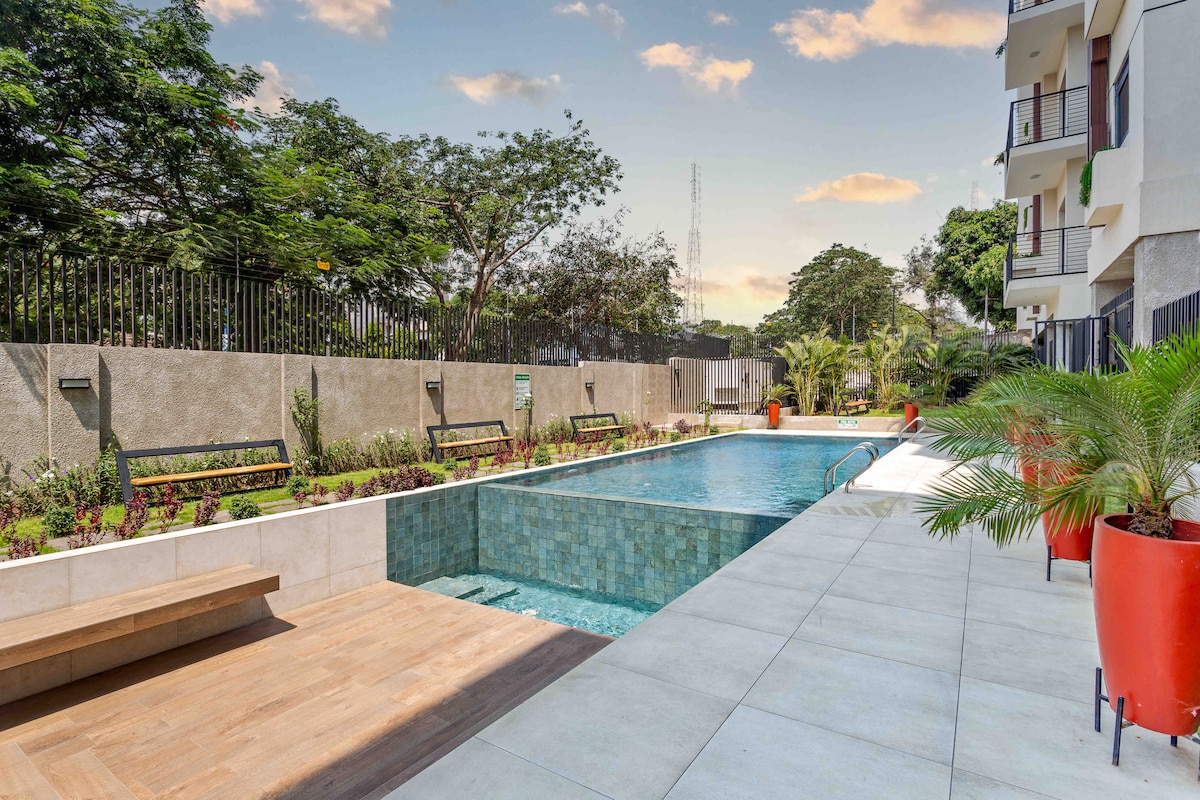 The image depicts a serene swimming pool surrounded by lush greenery and vibrant potted plants. Benches line the pool area, and the clean, modern design offers a relaxing space enhanced by natural light and soft reflections on the water.
