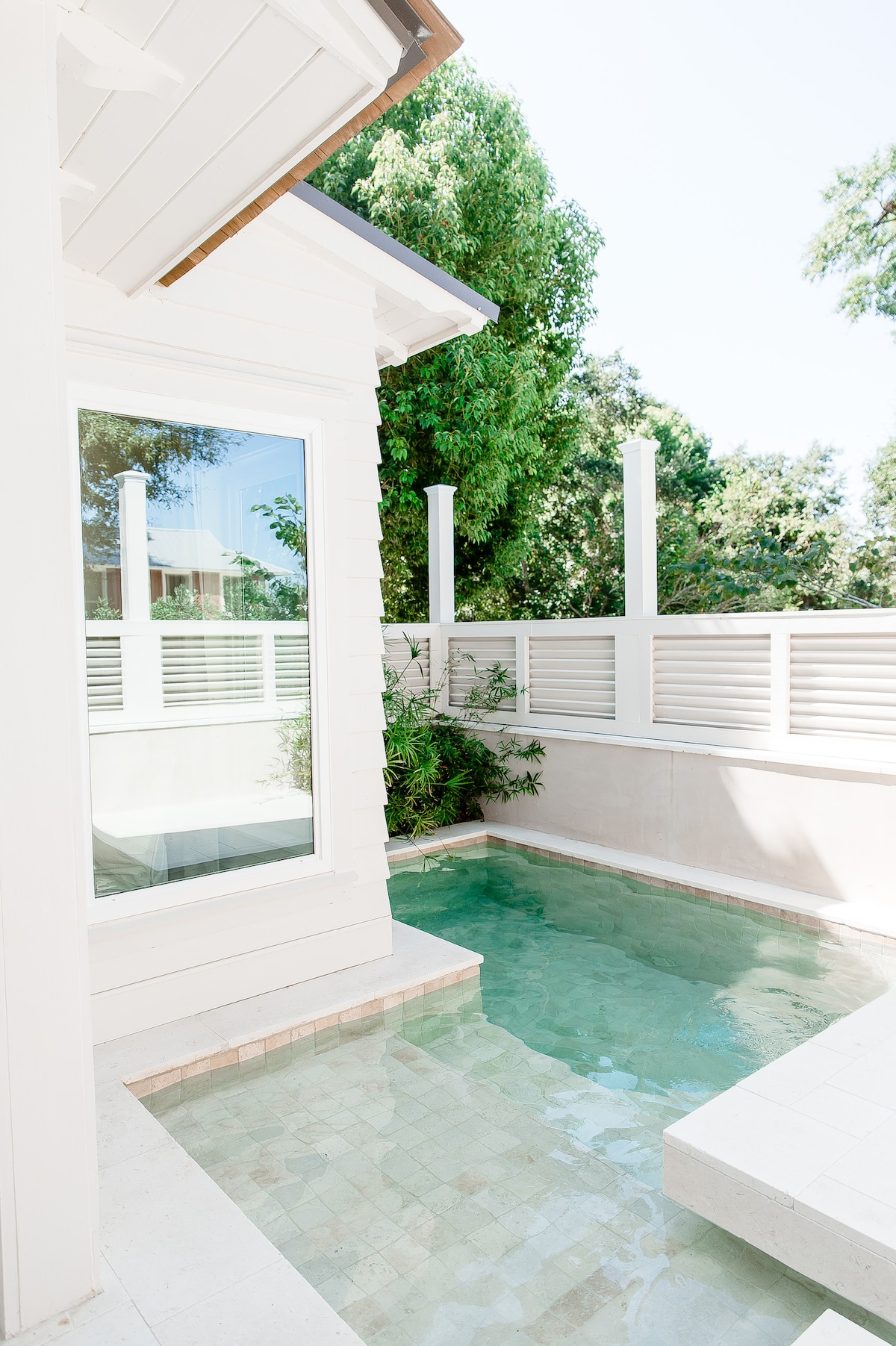 A spa pool is situated next to a cottage, featuring clear water reflecting sunlight. White wooden panels provide privacy, while greenery borders the area, enhancing the tranquil atmosphere. The pool's tiled edge leads to a small, integrated seating space for relaxation.