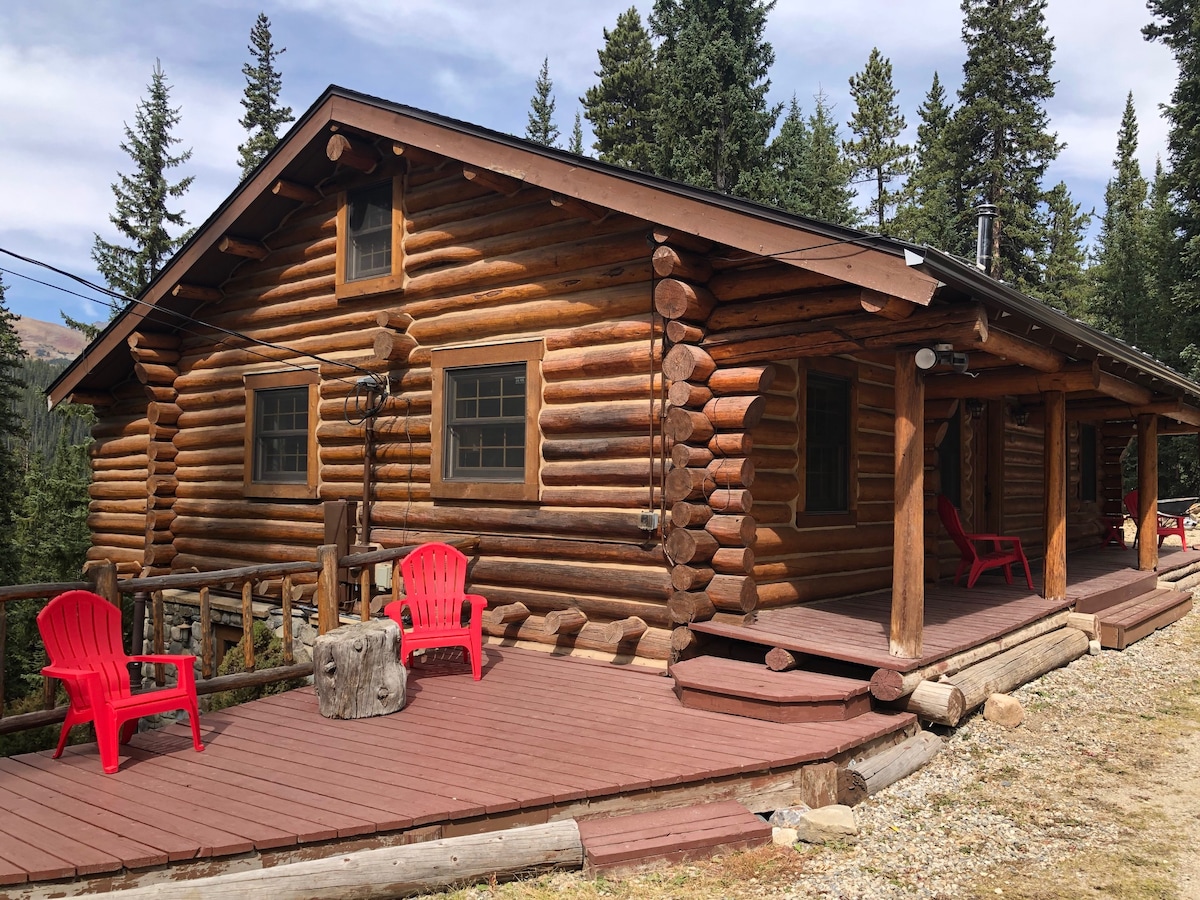 A rustic log home is situated among tall evergreen trees, featuring a covered porch with two red chairs placed on a wooden deck. Natural wooden elements are incorporated in the structure, creating a harmonious blend with the surrounding landscape.